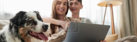 niedlichen australischen Schäferhund in der Nähe fröhlich lgbt Paar lächelnd, während sie zusammen sitzen und umarmen einander neben Laptop auf verschwommenem Hintergrund im Wohnzimmer, Banner 