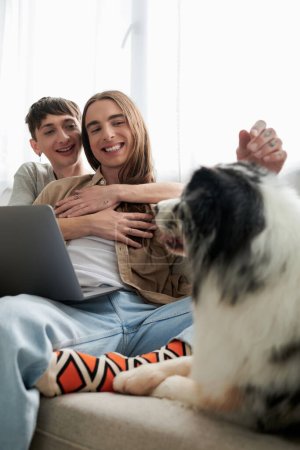 fröhliche homosexuelle Partner in lässiger Kleidung lächeln, während sie sich umarmen und zusammen neben Laptop sitzen und netten pelzigen Freund neben sich auf der Couch im modernen Wohnzimmer ausruhen