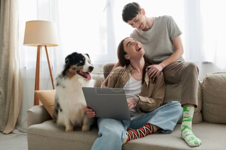 Australian shepherd dog resting on comfortable couch near cheerful gay partners in casual clothes smiling while hugging each other and sitting together with laptop 