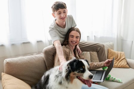 fröhlicher schwuler Mann kuschelt australischen Schäferhund, während er neben einem glücklichen Freund mit langen Haaren sitzt und Laptop hält, während er von zu Hause aus im Wohnzimmer arbeitet 