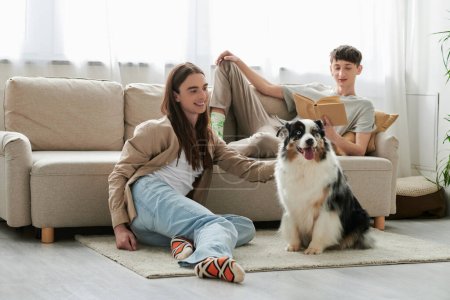 glücklicher schwuler Mann liest Buch und liegt auf bequemer Couch, während sein Freund mit langen Haaren in seiner Freizeit zu Hause mit einem australischen Schäferhund spielt 