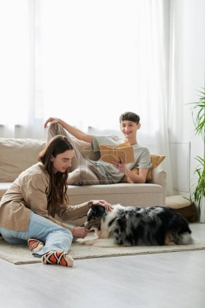 Foto de Alegre gay hombre leyendo libro y descansando en cómodo sofá mientras su novio con largo pelo jugando con australiano pastor perro en moderno apartamento - Imagen libre de derechos