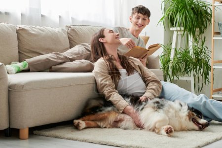 fröhlicher schwuler Mann mit Buch auf bequemer Couch, während sein Freund mit langen Haaren auf Teppich neben australischem Schäferhund in moderner Wohnung sitzt 