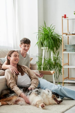 fröhlicher schwuler Mann hält Buch in der Hand und ruht sich auf bequemer Couch aus, während er seinen Freund mit langen Haaren umarmt, der neben dem australischen Schäferhund in der modernen Wohnung auf dem Teppich sitzt 