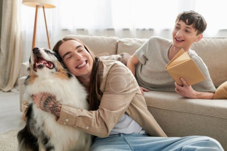 glücklicher schwuler Mann mit Tätowierung lächelt, während er den australischen Schäferhund neben einem fröhlichen schwulen Mann umarmt, der ein Buch in der Hand hält und sich auf einer bequemen Couch im Wohnzimmer ausruht 