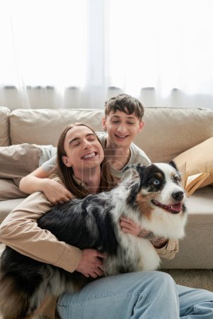fröhliches lgbt-Paar in lässiger Kleidung, das neben einer bequemen Couch sitzt und zusammen lächelt, während es den australischen Schäferhund im modernen Wohnzimmer schmust 