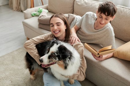 fröhlicher schwuler Mann liest Buch und ruht auf bequemem Sofa neben seinem Freund mit langen Haaren auf Teppich sitzend mit australischem Schäferhund in moderner Wohnung 