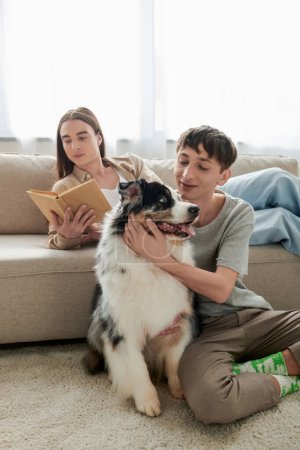 junger schwuler Mann mit langen Haaren liest Buch und ruht auf bequemem Sofa neben seinem Freund mit Tätowierung sitzt auf Teppich mit australischem Schäferhund in moderner Wohnung 