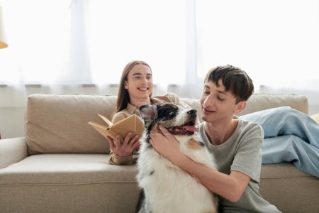 glücklicher junger schwuler Mann mit langen Haaren, Buch in der Hand und auf bequemem Sofa neben seinem Freund mit tätowiertem kuschelndem australischem Schäferhund in moderner Wohnung 