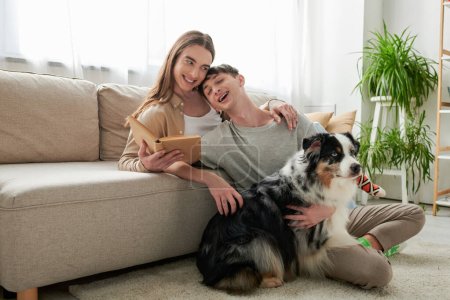 glücklicher schwuler Mann mit langen Haaren hält Buch in der Hand und umarmt seinen tätowierten und fröhlichen Freund neben dem australischen Schäferhund im modernen Wohnzimmer zu Hause 