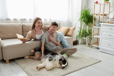 fröhlicher Mann auf Teppich sitzend und schmusend australischer Schäferhund und anrührende Hand seines schwulen Partners mit langen Haaren hält Buch im modernen Wohnzimmer 