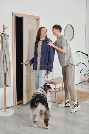 pleased gay couple in casual outfits standing in modern hallway next to coat rack and holding leash with Australian shepherd dog and smiling together at home 