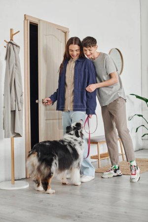 glückliches schwules Paar in lässigen Outfits, das im modernen Flur neben der Garderobe steht und den australischen Schäferhund mit Leine betrachtet und zu Hause zusammen lächelt 