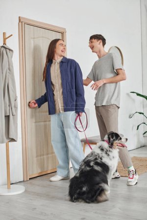 Photo for Happy gay man looking at boyfriend with long hair standing in modern hallway next to coat rack and holding leash with Australian shepherd dog while smiling together at home - Royalty Free Image