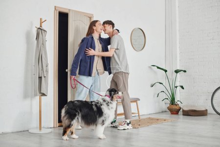 glückliches schwules Paar in lässigem Outfit, das im Flur neben der Garderobe steht und sich umarmt und in der Nähe des australischen Schäferhundes an der Leine hält, während es in der modernen Wohnung zusammen lächelt