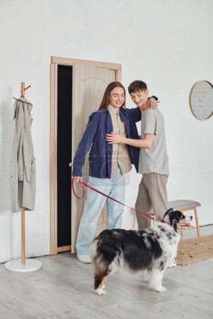 glückliches schwules Paar in lässigem Outfit, das im Flur neben der Garderobe steht und sich umarmt und in der Nähe eines australischen Schäferhundes an der Leine hält und in einer modernen Wohnung zusammen lächelt