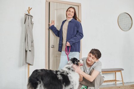 glücklicher schwuler Mann lächelt, während er liebenswert australischen Schäferhund neben fröhlichen Freund mit langen Haaren an der Leine hält, während er in der Nähe der Tür im modernen Flur steht 