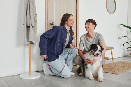 cheerful lgbt couple in casual outfits smiling while kneeling together and cuddling cute Australian shepherd dog next to door and coat rack in hallway of modern apartment 