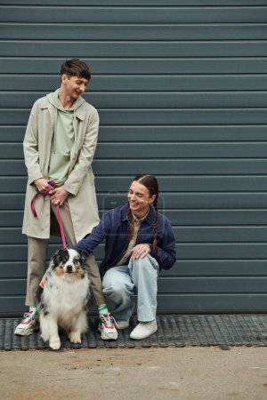 cheerful gay man with pigtails cuddling Australian shepherd dog next to smiling boyfriend in coat holding leash near garage door outside on street