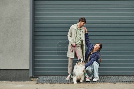 happy and young gay man with pigtails cuddling Australian shepherd dog next to smiling boyfriend in coat holding leash near garage door outside on street