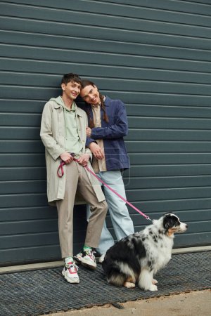 cheerful gay man in casual outfit holding leash of Australian shepherd dog and standing next to smiling boyfriend with pigtails near garage door outside on street