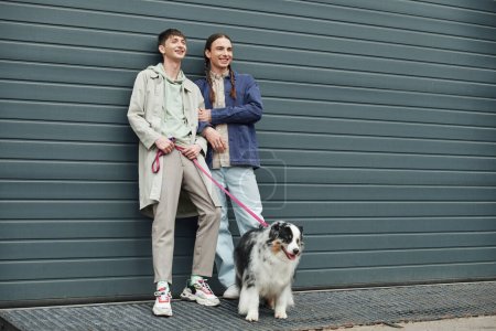 Photo for Tattooed and cheerful gay man in casual outfit holding leash of Australian shepherd dog and standing next to positive boyfriend with pigtails hairstyle near garage door outside on street - Royalty Free Image