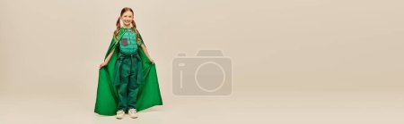 niña sonriente en traje de superhéroe verde con capa usando pantalones y camiseta y de pie mientras se celebra la fiesta del día de protección del niño sobre fondo gris, pancarta 