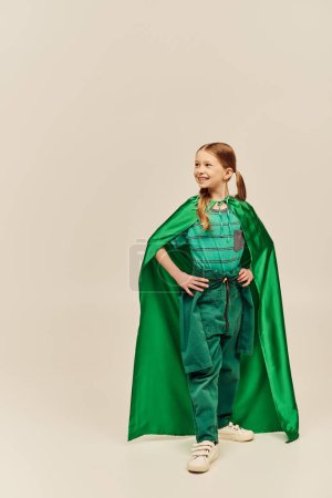 smiling girl in green superhero costume with cloak wearing pants and t-shirt and standing with hands on hips while celebrating International Children Day on grey background 