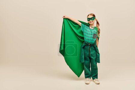 niño feliz en traje de superhéroe y máscara en la cara sosteniendo capa verde, usando pantalones y camiseta y de pie mientras se celebra la fiesta del día de protección del niño sobre fondo gris 