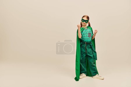 fille puissante en costume de super-héros vert avec manteau et masque sur le visage, portant un pantalon et un t-shirt et debout avec des poings serrés tout en célébrant les vacances de la journée de protection de l'enfance sur fond gris 