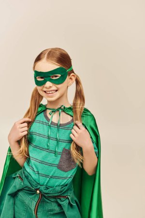 lächelndes Mädchen in grünem Superheldenkostüm mit Mantel und Maske im Gesicht, mit Zwillingsschwanzfrisur, die ihr Haar berührt, während sie den Internationalen Kindertag auf grauem Hintergrund feiert 