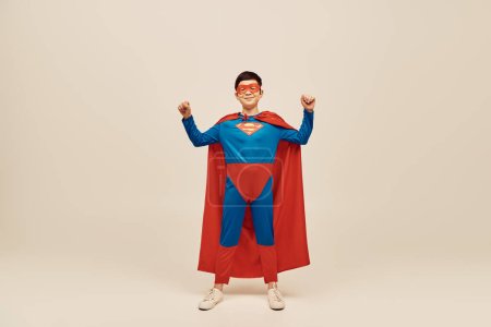 Foto de Feliz asiático chico en rojo y azul superhéroe traje con capa y máscara en la cara mostrando fuerza gesto mientras celebrando feliz día de los niños en gris fondo - Imagen libre de derechos