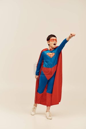 azjatycki chłopiec w stroju superbohatera z płaszczem i maską na twarzy krzyczy pokazując jednocześnie gest siły stojąc z wyciągniętą ręką na szarym tle, koncepcja dzień ochrony dziecka 