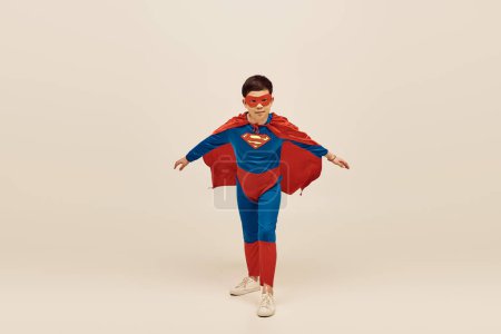 valiente asiático chico en rojo y azul superhéroe traje con capa y máscara en la cara celebrando el Día Internacional de la Protección de los Niños sobre fondo gris 