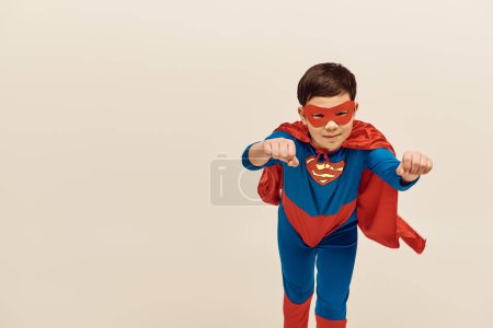 Mutiger asiatischer Junge im Superheldenkostüm mit Mantel und Maske, der mit ausgestreckten Händen und geballten Fäusten den Internationalen Tag zum Schutz von Kindern auf grauem Grund feiert 