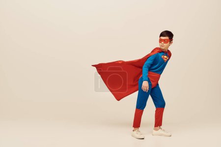 longitud completa de valiente asiático chico en traje de superhéroe con capa y máscara mirando hacia otro lado y de pie contra el viento durante el Día Mundial de la Protección del Niño sobre fondo gris 