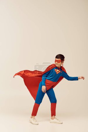 in voller Länge glücklicher asiatischer Junge im Superheldenkostüm mit Mantel und Maske lächelt beim Wegschauen und steht gegen den Wind während des Internationalen Kinderferientages auf grauem Hintergrund 