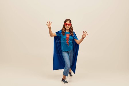 fröhliches Mädchen im Superheldenkostüm mit blauem Mantel und roter Maske im Gesicht, mit Handbewegung, stehend in Jeans und T-Shirt beim Kinderschutztag auf grauem Hintergrund 