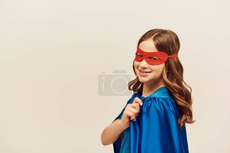 happy girl in superhero costume with blue cloak and red mask on face looking at camera and smiling while celebrating International children's day on grey background 