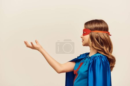 Téléchargez les photos : Vue latérale de fille heureuse en costume de super-héros avec manteau bleu et masque rouge sur le visage, debout avec la main tendue pendant sur fond gris en studio, concept de Journée mondiale de la protection de l'enfance - en image libre de droit