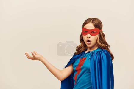 Superhéros choqué fille en costume avec manteau bleu et masque rouge debout avec la main tendue et la bouche ouverte pendant la Journée internationale de la protection de l'enfance sur fond gris en studio 