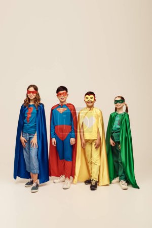 niños preadolescentes multiétnicos felices en trajes coloridos de superhéroes con capas y máscaras de pie juntos mientras celebran las vacaciones del día de protección infantil sobre fondo gris en el estudio 