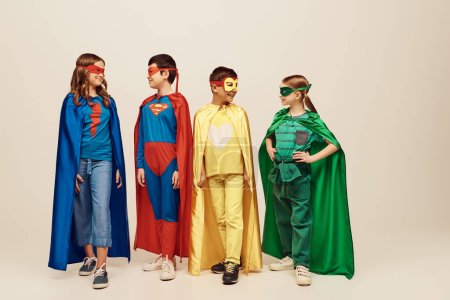 fröhliche interrassische Vorschulkinder in bunten Superheldenkostümen mit Mänteln und Masken, die auf grauem Hintergrund im Studio stehen und einander anschauen, Weltkinderschutztag 
