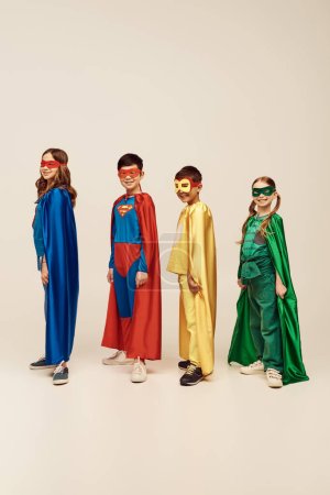 cheerful interracial kids in colorful superhero costumes with cloaks and masks standing together and looking at camera while celebrating Child protection day holiday on grey background in studio 