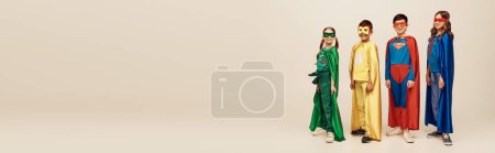 happy interracial preteen kids in colorful costumes with cloaks and masks standing together while celebrating Child protection day holiday on grey background in studio, banner 