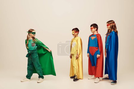 fröhliche interrassische Preteen Kids in bunten Superheldenkostümen betrachten Mädchen in grünem Mantel und Maske, während sie den Kinderschutztag auf grauem Hintergrund im Studio feiern 