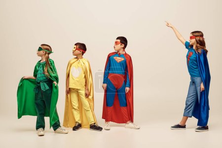 glückliches Mädchen in Superheldenmantel und Maske, das in der Nähe von Freunden in Kostümen steht und wegschaut, während es im Studio den Kinderschutztag auf grauem Hintergrund feiert 