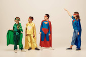 happy preteen girl standing in superhero cloak and mask, pointing away near interracial friends in costumes looking away while celebrating Child protection day on grey background in studio  Sweatshirt #655801534