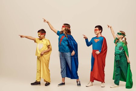 Positive multikulturelle Kinder in bunten Superheldenkostümen mit Mänteln und Masken, die mit den Fingern zeigen, während sie vor grauem Hintergrund im Studio wegschauen, Internationaler Kinderschutztag