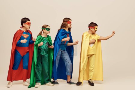 enfants multiculturels courageux en costumes colorés avec des manteaux et des masques debout avec des poings serrés ensemble sur fond gris en studio, concept de Journée de la protection de l'enfance